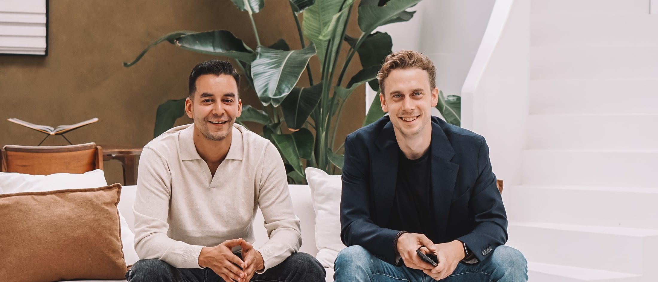Mo en Paul zitten naast elkaar op een bank en kijken lachend in de camera